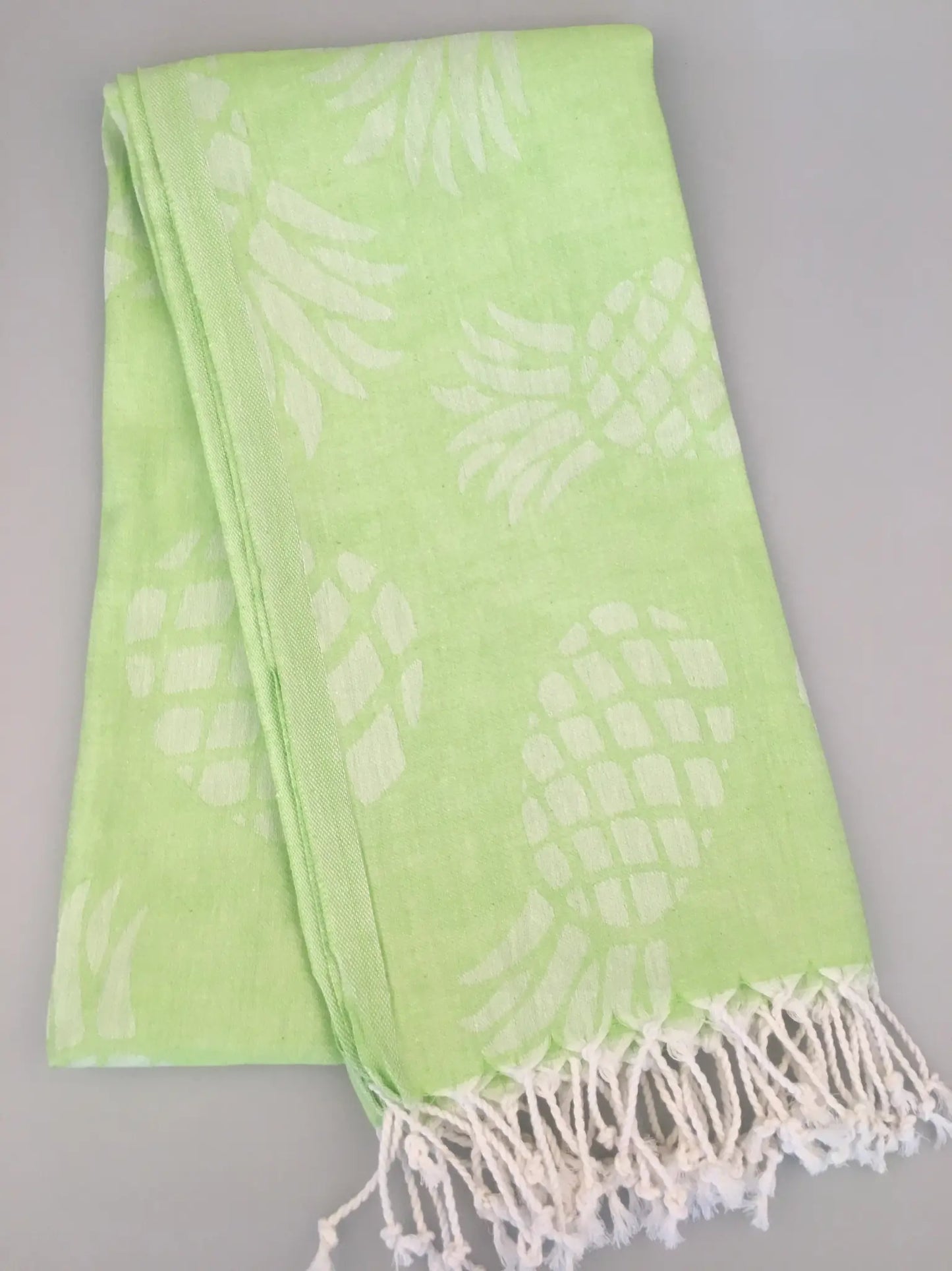 Light green Turkish towel with white pineapple jacquard pattern and fringed edges, folded view on a gray background.