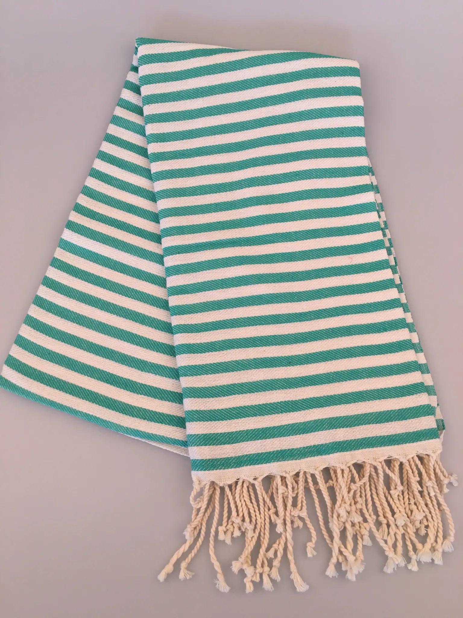 Turquoise and white striped Turkish towel with fringe edges, folded at an angle on a light background.