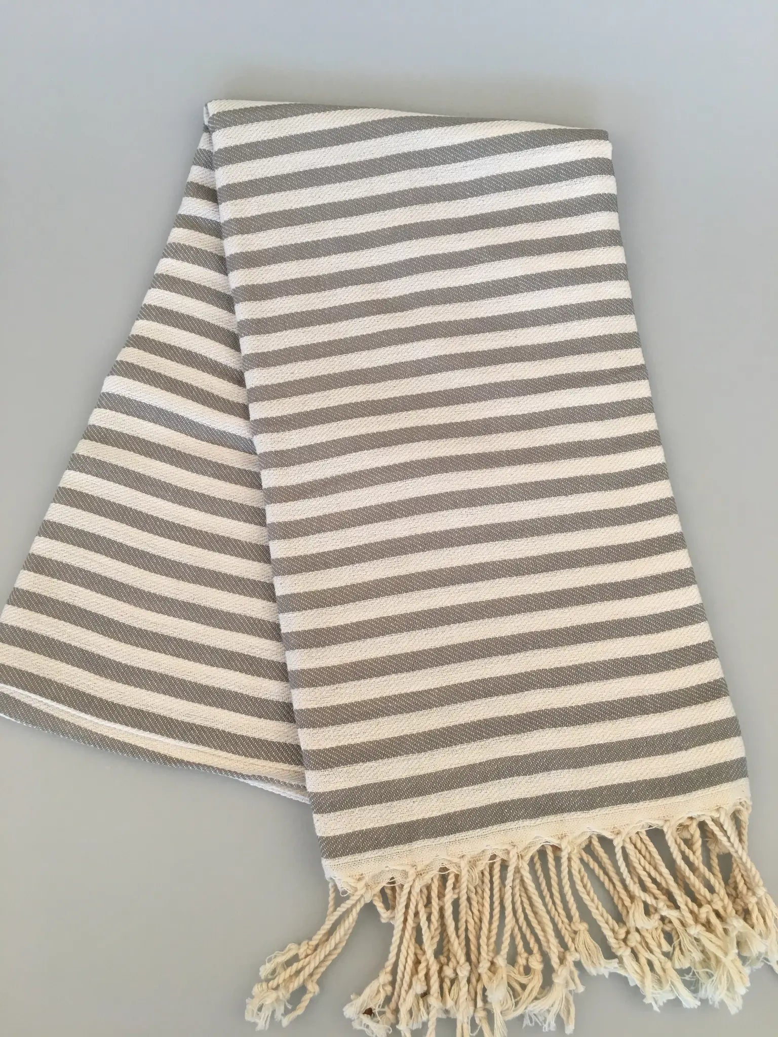 Gray and cream striped Turkish towel with fringed edges, neatly folded on a light gray background.