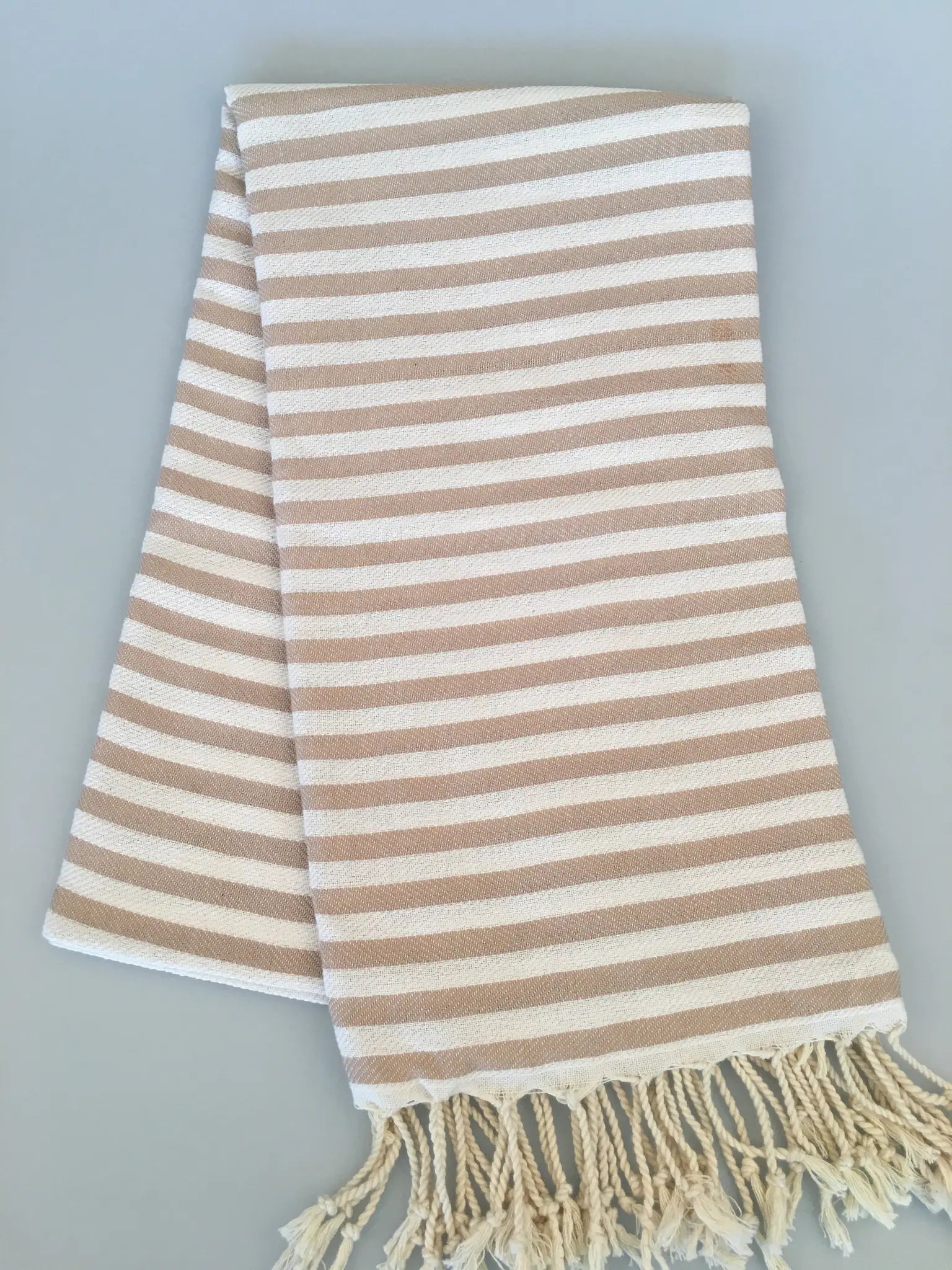 "Beige and cream striped Turkish towel with fringed edges, folded flat on a light gray background."