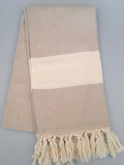 Beige Turkish towel peshtemal with herringbone pattern, cream stripe, and fringed edges, laid flat on a gray background.