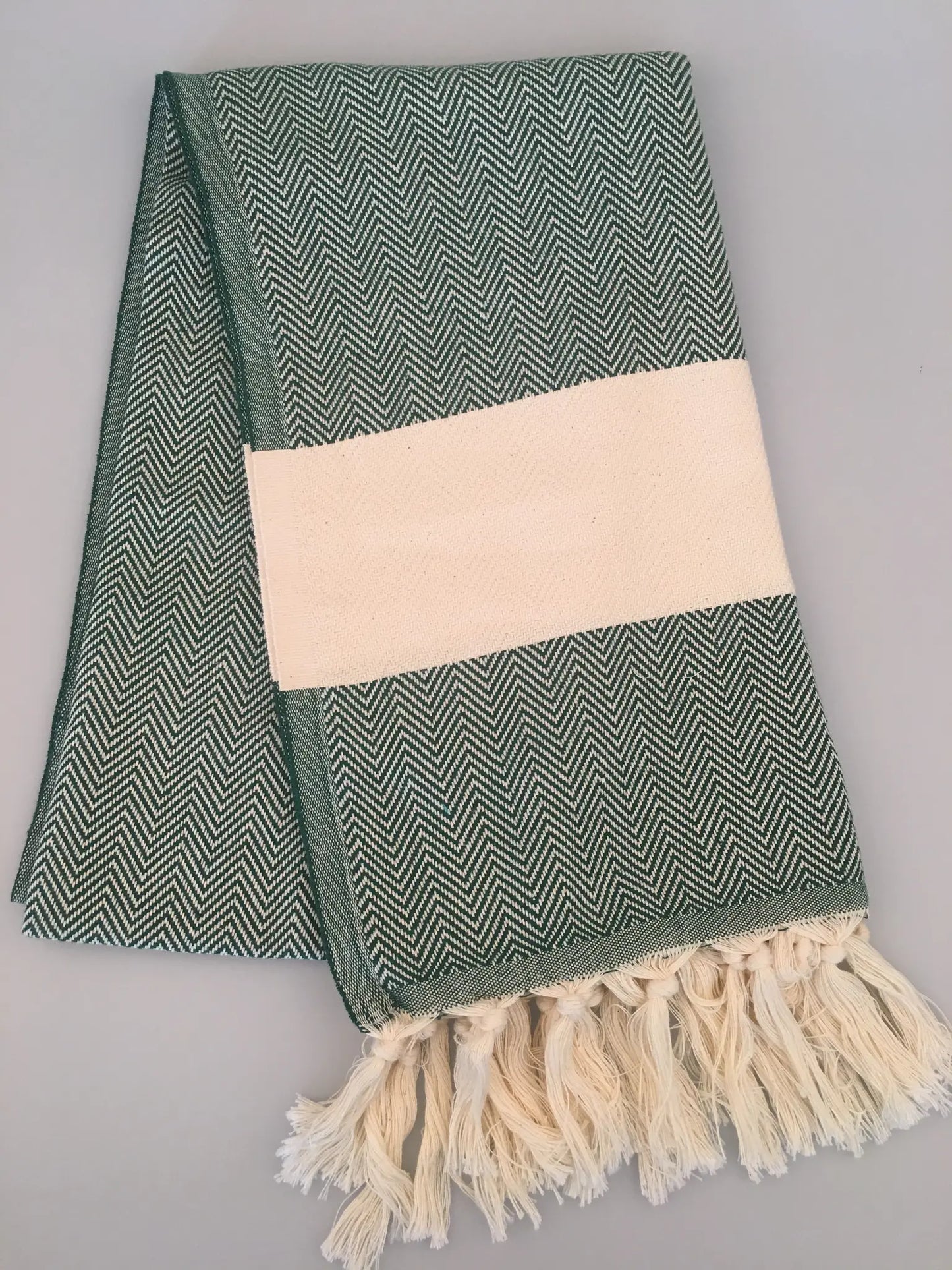 Green and white herringbone-patterned Turkish towel with fringed edges and a solid cream band, laid flat on a light backgroun