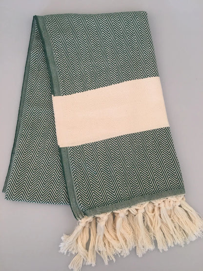 Green and white herringbone-patterned Turkish towel with fringed edges and a solid cream band, laid flat on a light backgroun