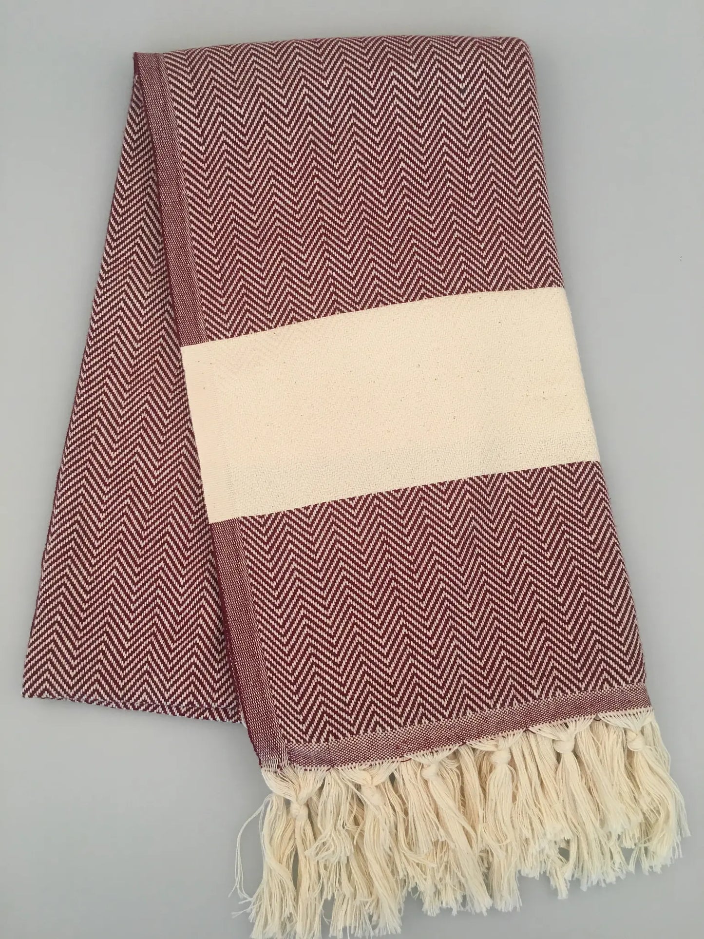 Burgundy Turkish towel with a herringbone pattern, cream stripe, and fringed edges, folded on a light gray background.