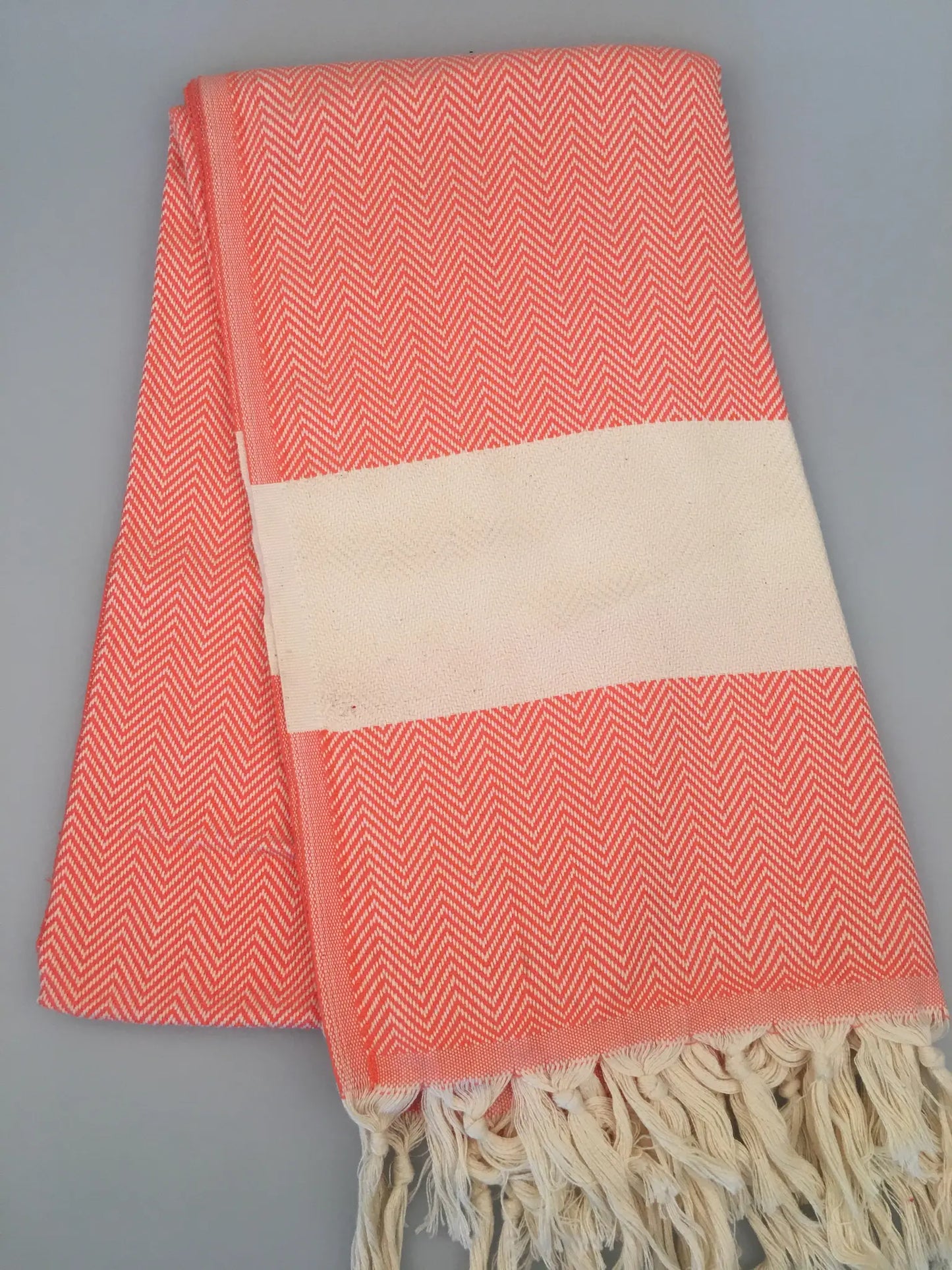 Coral and white Turkish towel with herringbone pattern, featuring a cream-colored band and fringed edges, laid flat.