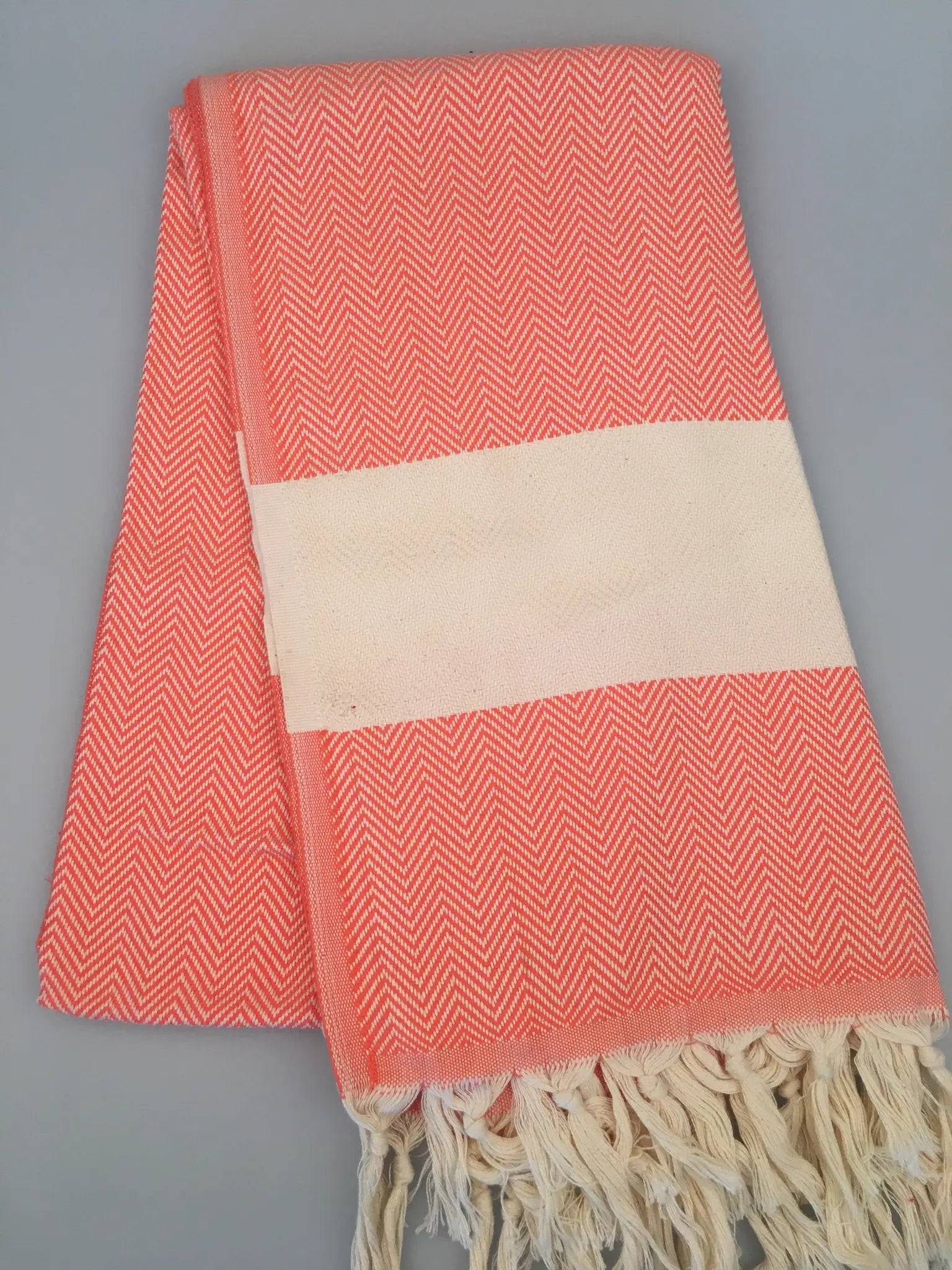 Coral and white Turkish towel with herringbone pattern, featuring a cream-colored band and fringed edges, laid flat.