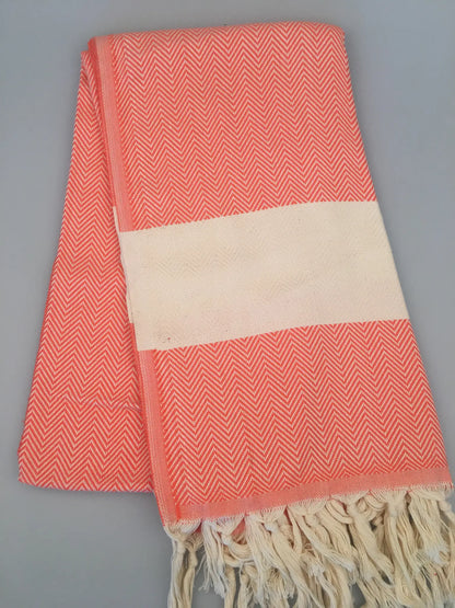 Coral and white Turkish towel with herringbone pattern, featuring a cream-colored band and fringed edges, laid flat.