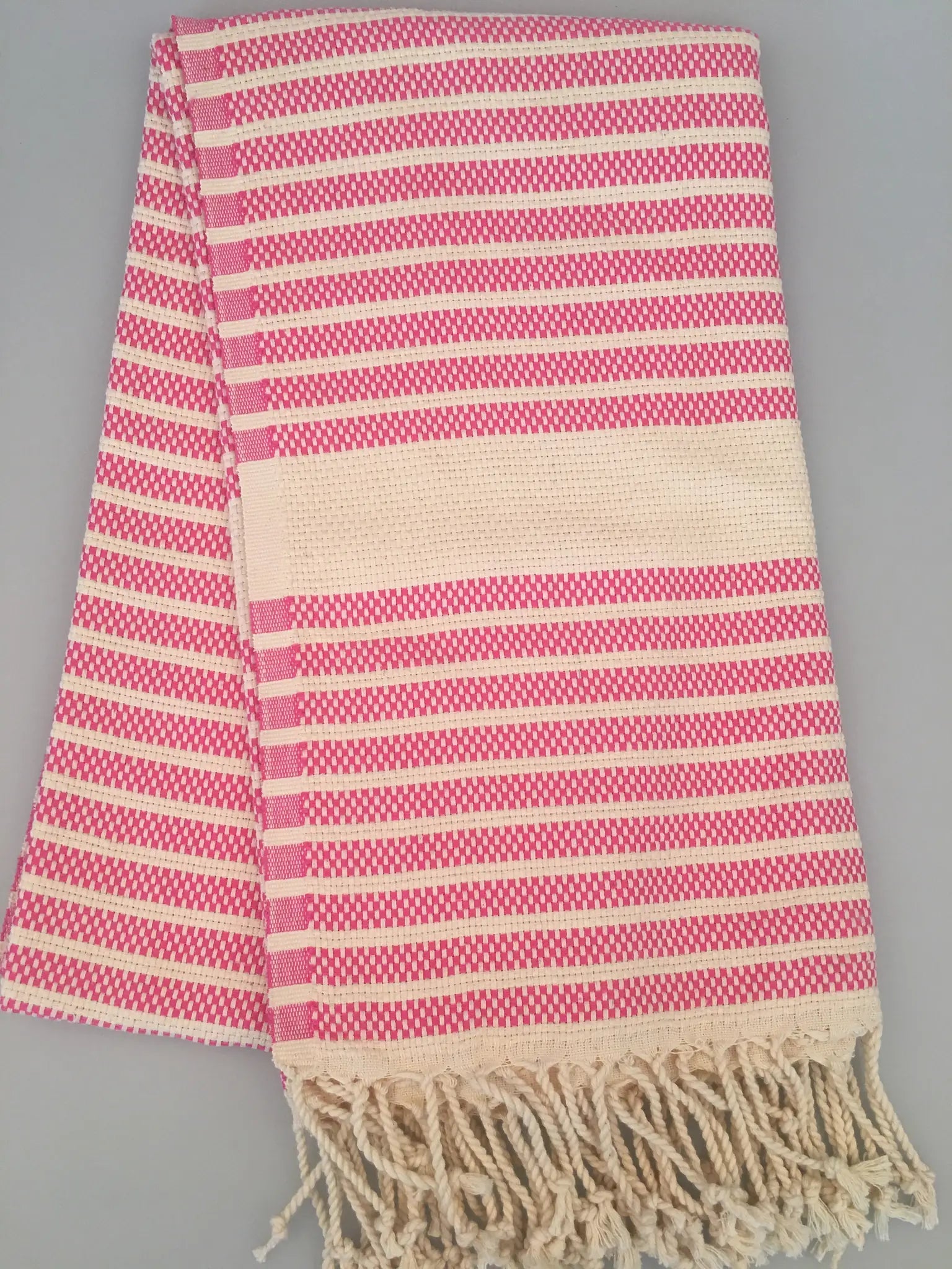 "Pink and cream striped Turkish towel with fringed edges, folded on a gray background."