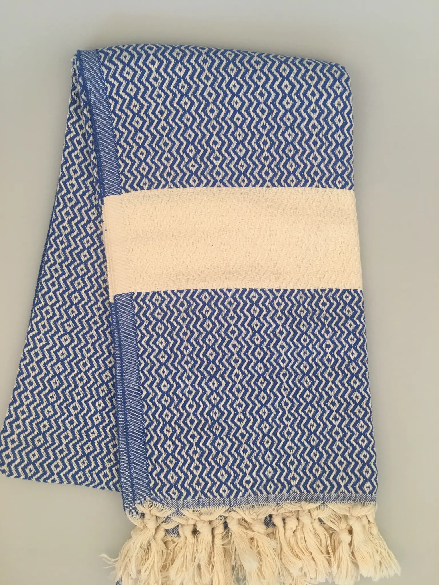 "Blue and white patterned Turkish towel with fringed edges and a solid cream band, neatly folded on a light surface."