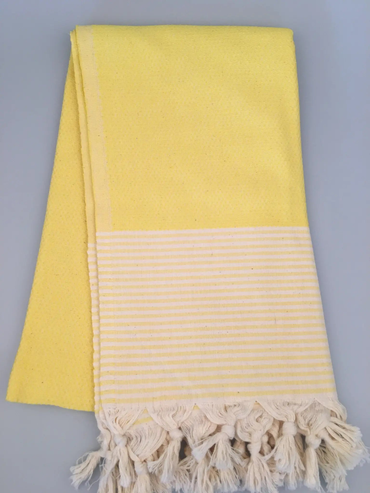 "Yellow Turkish towel with textured weave and striped bottom panel, fringed edges, folded neatly."