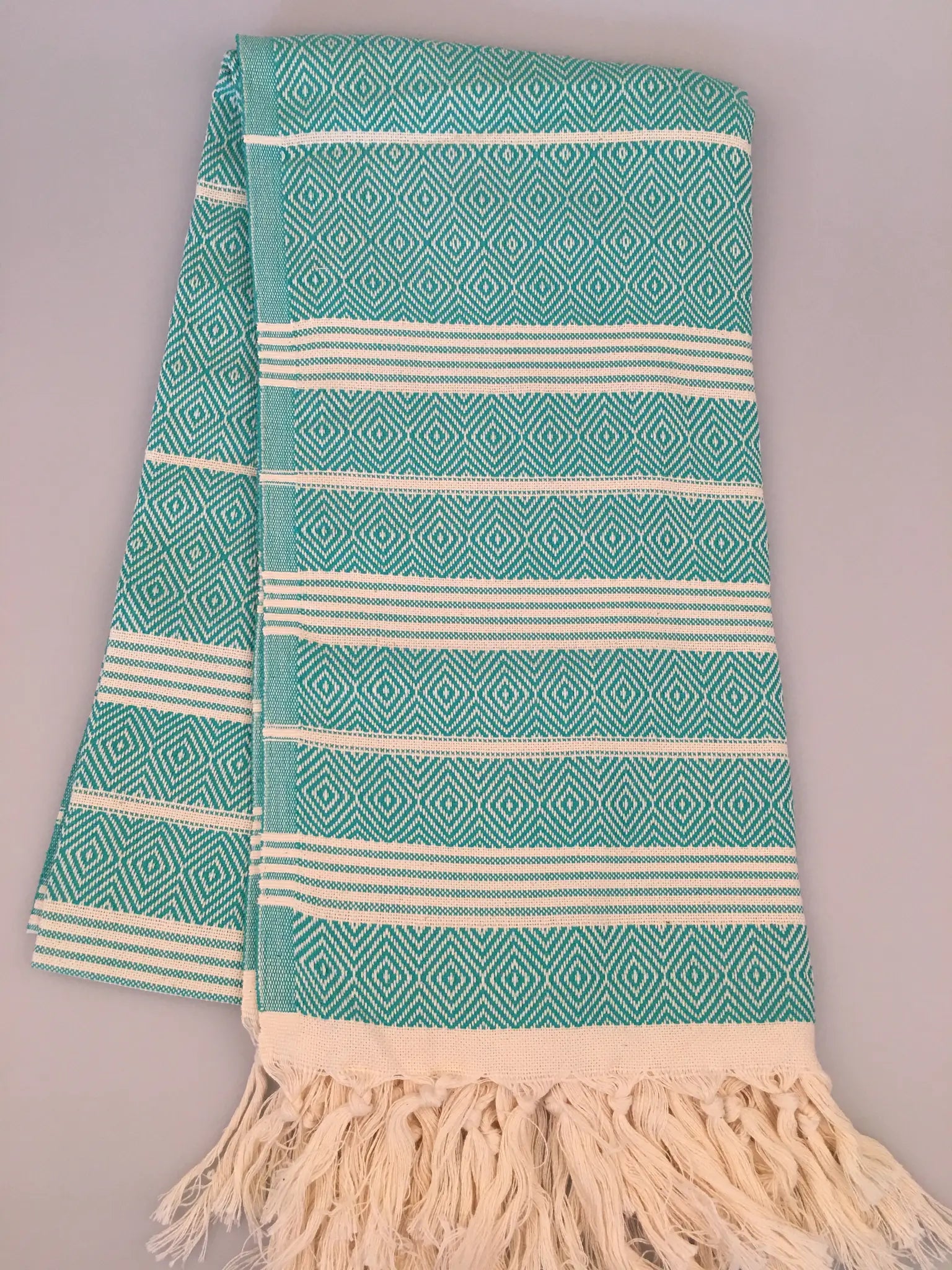 "Turquoise Turkish towel with diamond patterns and white stripes, fringed edges, laid flat on a neutral background."