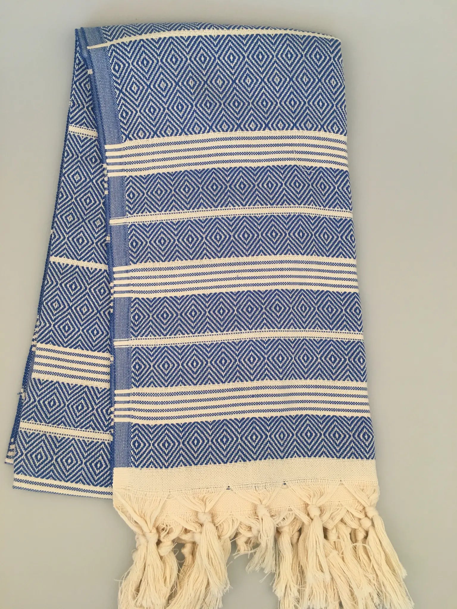 "Lycia Xanthos Turkish towel peshtemal in blue and white diamond pattern with fringed edges, folded on a light surface."