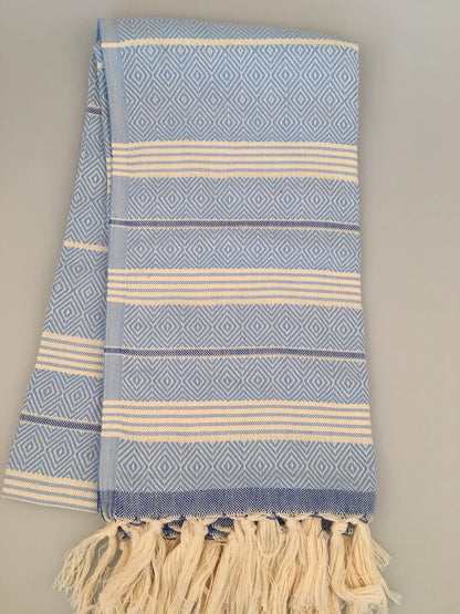 Light blue Turkish towel with diamond patterns, white stripes, and fringed edges, laid flat on a neutral background.