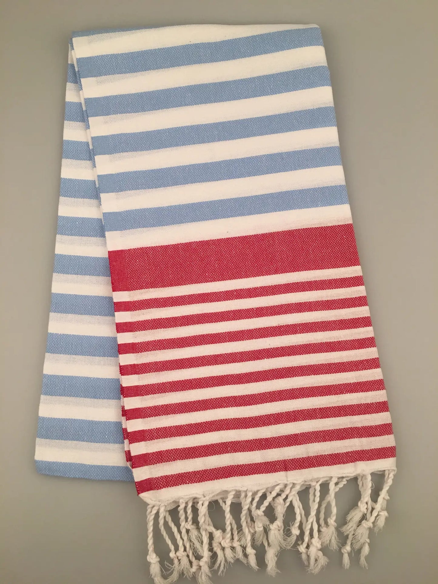 "Striped peshtemal towel in light blue and white with a red striped section, fringed ends, laid flat."