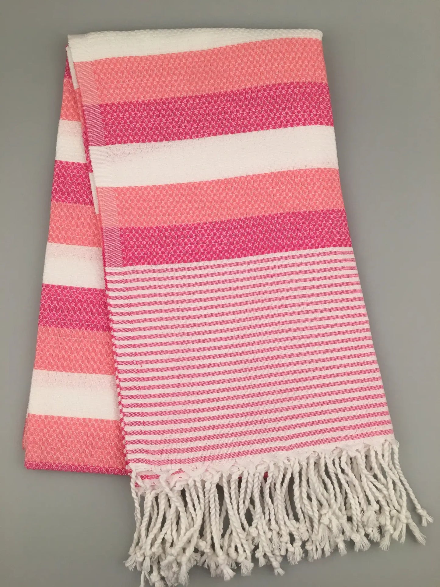Pink and white Turkish towel with striped pattern and fringed edges, folded on a light gray background.