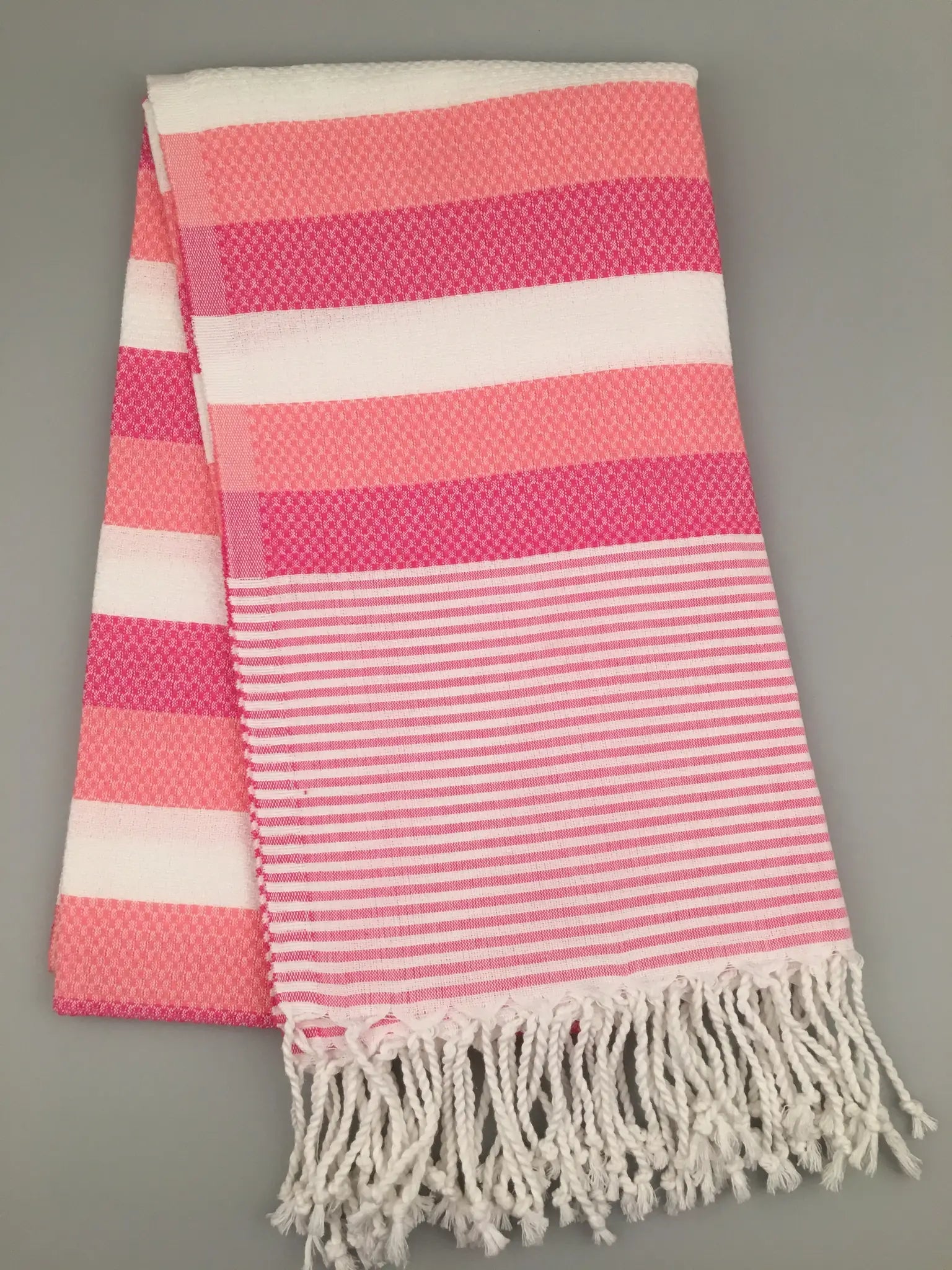 Pink and white Turkish towel with striped pattern and fringed edges, folded on a light gray background.