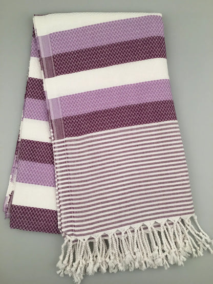 "Purple and white striped Turkish towel with textured patterns and fringed edges, laid flat on a gray background."