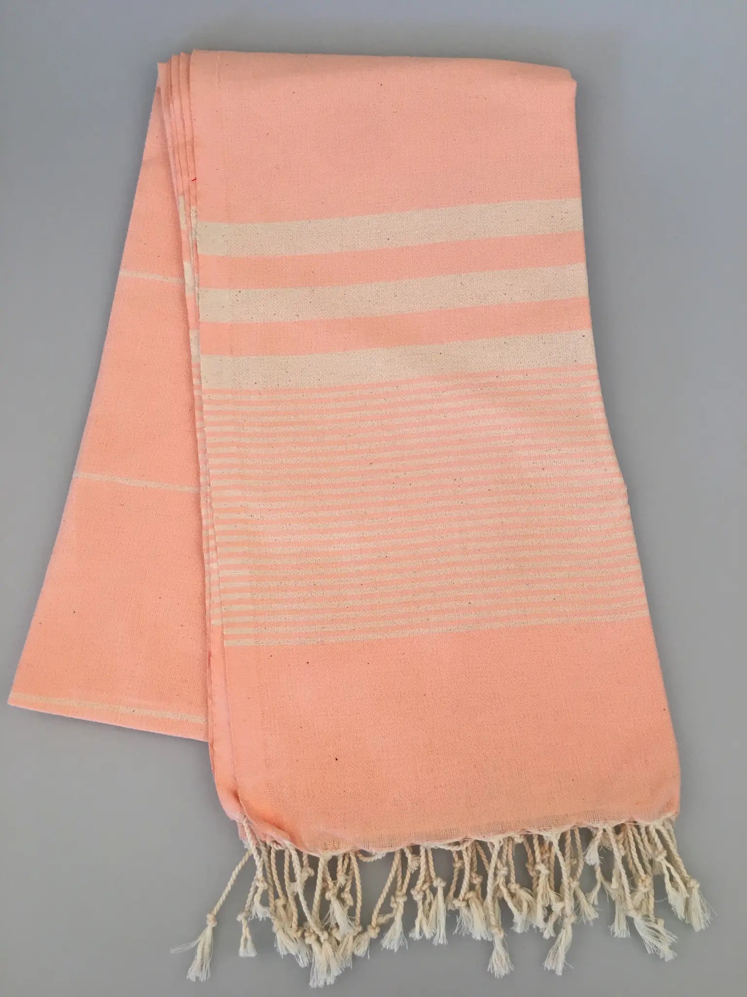 "Peach Turkish towel with cream stripes and tassels, folded on a light gray background."