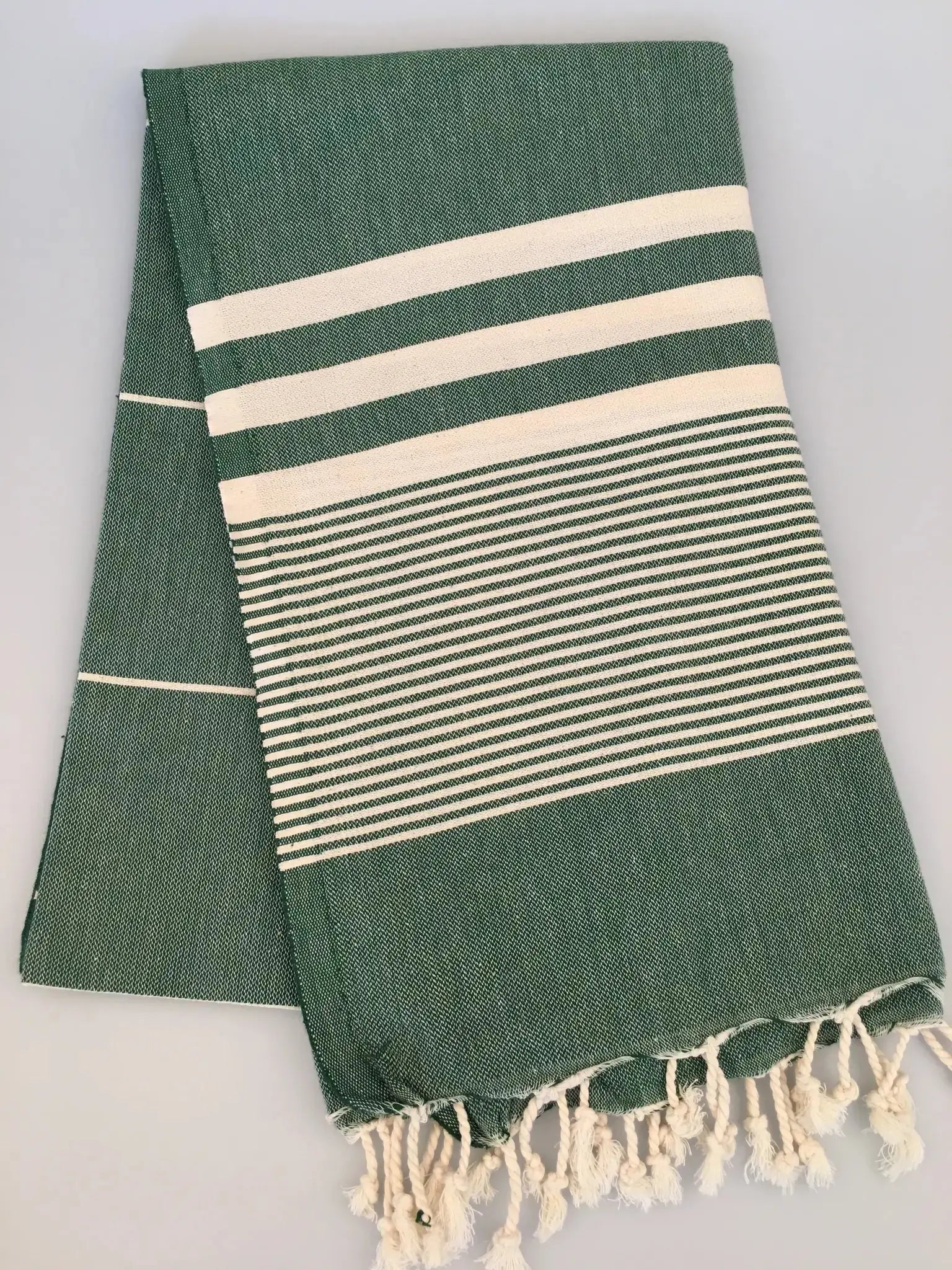 Green Turkish towel peshtemal with white stripes and fringed edges, folded neatly on a light background.
