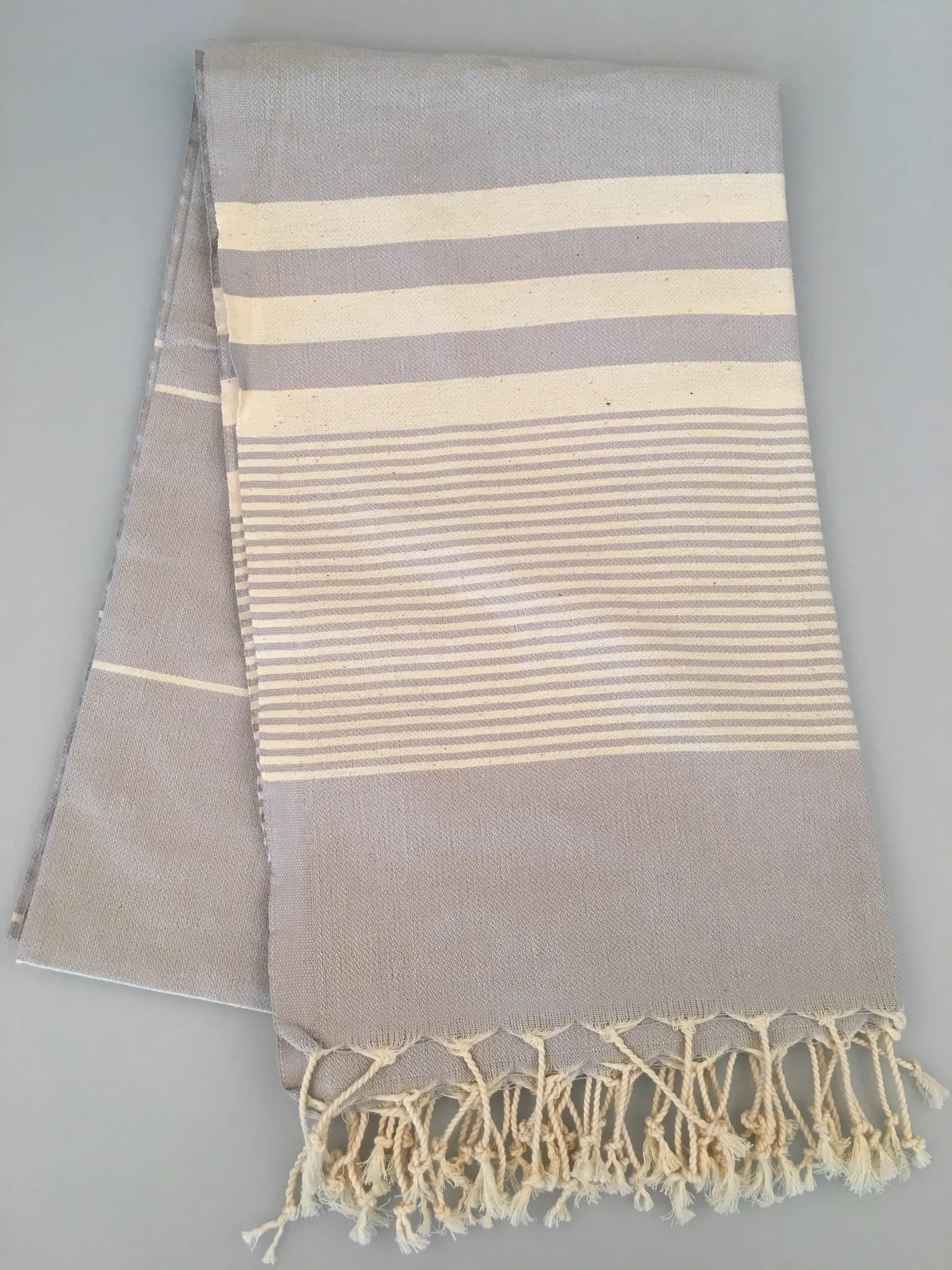 "Light gray Turkish towel with white stripes and fringed edges, folded view on a gray background."